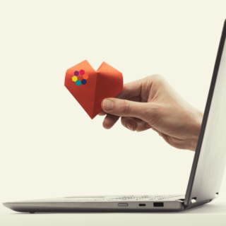 A hand offers a paper heart through a laptop screen.