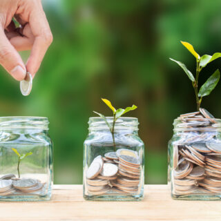 a hand dropping a coin into a glass jar with a plant growing out of it