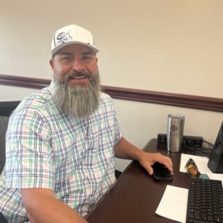 A man with a beard and ballcap smiles at the camera from his office desk.