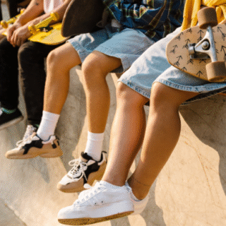 A group of teens sitting on the edge of a skate park ledge, holding skateboards. Only their legs and feet are visible, showing casual clothing and sneakers.
