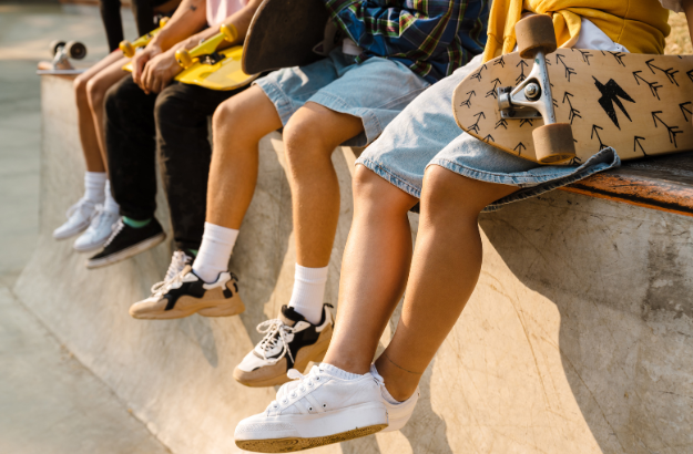 A group of teens sitting on the edge of a skate park ledge, holding skateboards. Only their legs and feet are visible, showing casual clothing and sneakers.